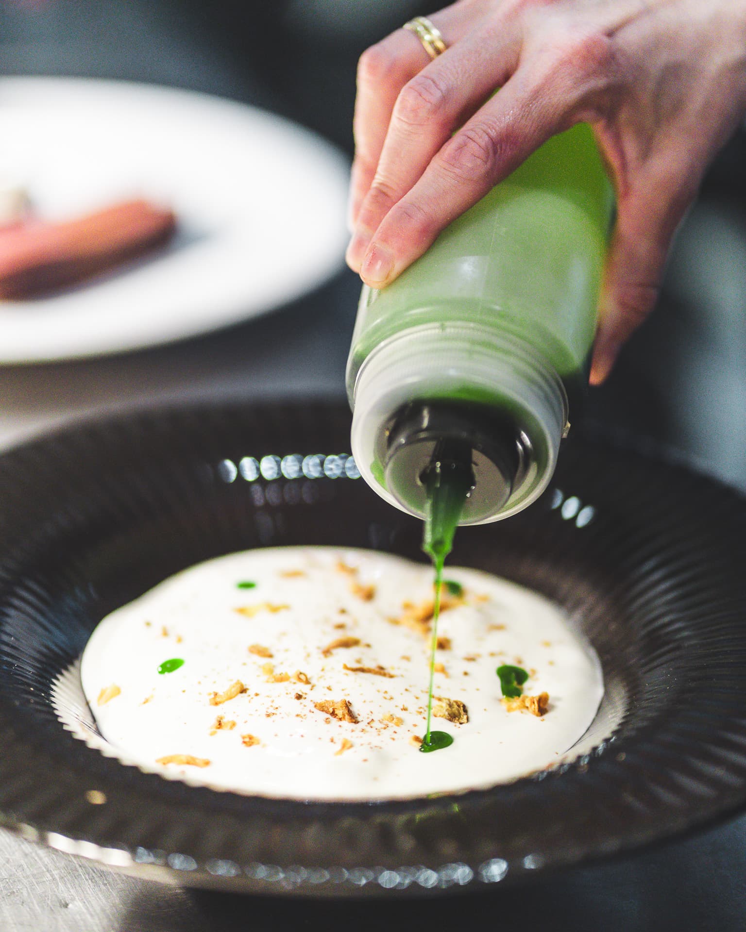 A chef's hand drizzling bright green herb oil over a cream-coloured base in a dark fluted bowl.